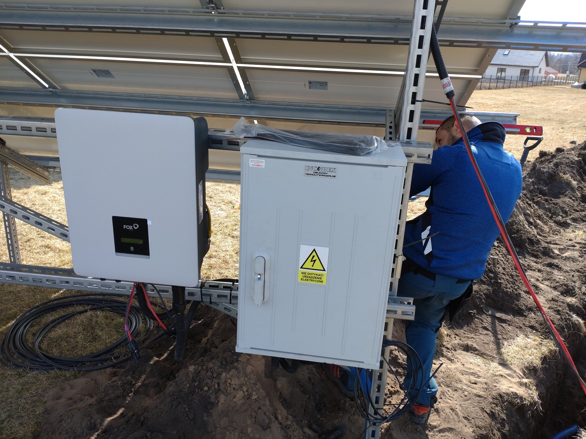 Instalacja paneli słonecznych: monter w niebieskiej bluzie poziomuje konstrukcję, widoczny inwerter FoxESS i skrzynka elektryczna z ostrzeżeniem o napięciu, w tle dom jednorodzinny.