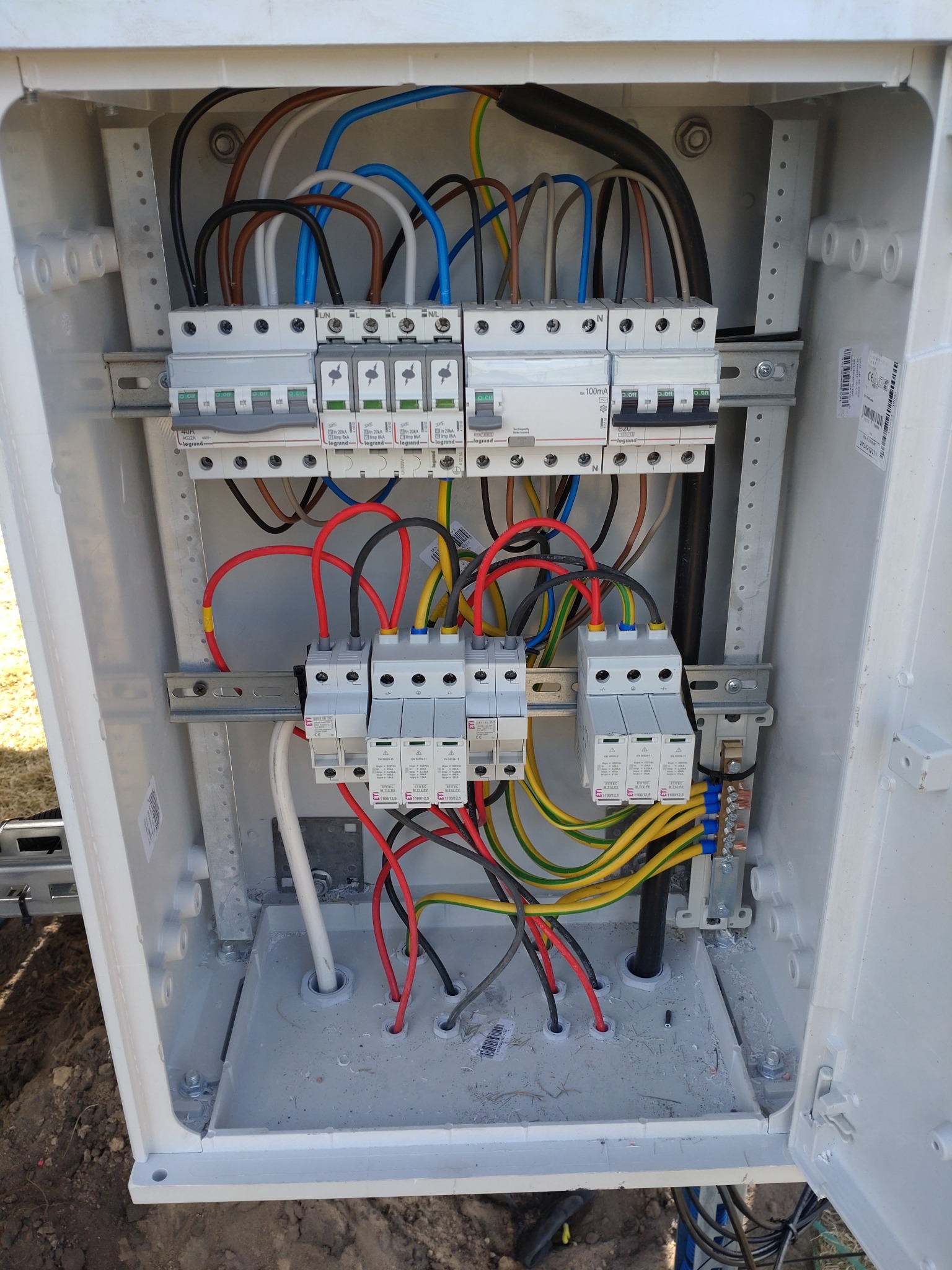 Otwarta skrzynka elektryczna z bezpiecznikami, przewodami w różnych kolorach i listwami zaciskowymi, instalacja fotowoltaiczna w trakcie montażu, widoczne ślady wiercenia.