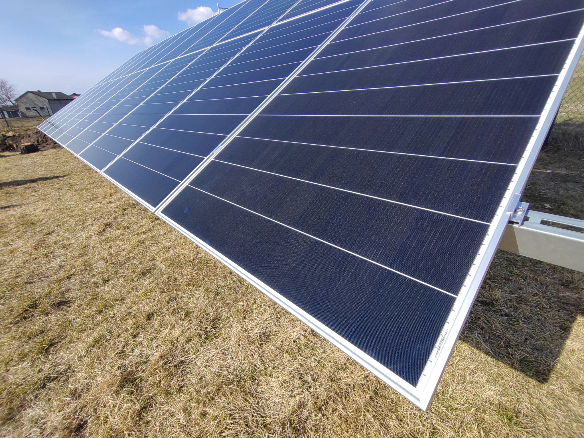 Nisko kątowe ujęcie paneli słonecznych zamontowanych na konstrukcji wsporczej, na tle wyschniętej trawy i błękitnego nieba z pojedynczymi chmurami.