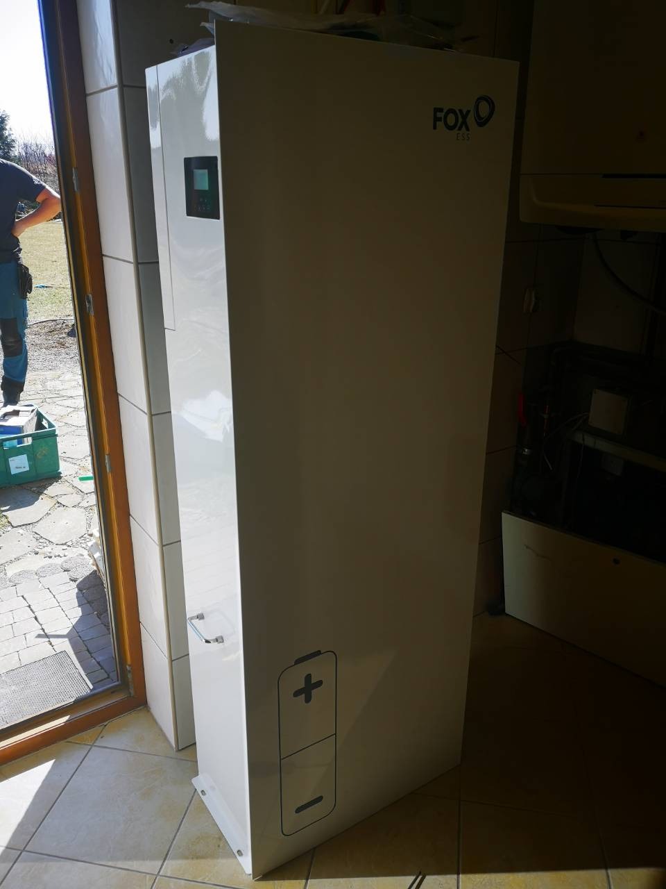 Biały, pionowy moduł magazynu energii Fox ESS z wyświetlaczem i symbolem baterii, umieszczony w pomieszczeniu z kafelkami, częściowo widoczny instalator przy otwartych drzwiach.