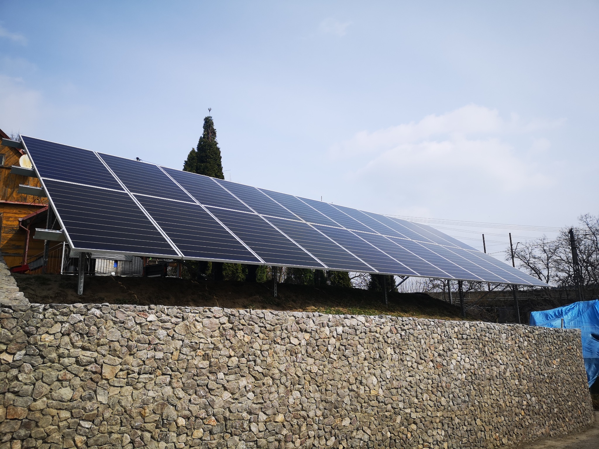 Instalacja paneli słonecznych na gruncie, widoczna zza kamiennego muru oporowego, w tle drzewa i błękitne niebo.