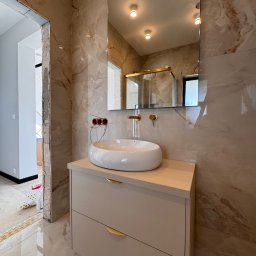 Mirage meble Adam Węgrzyński - Łazienka w trakcie wykończenia: szafka z umywalką nablatową, lustro, płytki imitujące marmur. Widoczne niedokończone obramowanie drzwi i wystające kable.