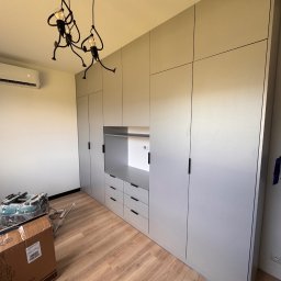 Mirage meble Adam Węgrzyński - Jasnoszara szafa na wymiar z szufladami i otwartą półką, oświetlona designerską lampą. Podłoga drewniana, widoczne narzędzia i kartony po montażu.