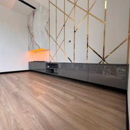 Mirage meble Adam Węgrzyński - Szara szafka RTV na wymiar z połyskiem, podświetlana od góry pomarańczowym światłem, na tle białej ściany z dekoracyjnymi, złotymi elementami.