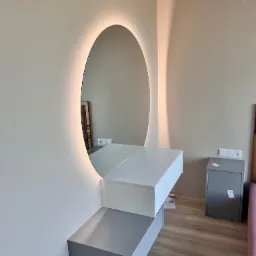 Nowoczesna toaletka z owalnym lustrem podświetlanym LED, zamontowana na ścianie w sypialni. Białe blaty tworzą minimalistyczny design, a drewniana podłoga dodaje ciepła.