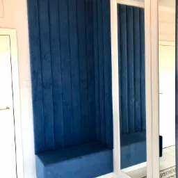 Narożna, tapicerowana na niebiesko zabudowa siedziska z pionowymi przeszyciami, wkomponowana w narożnik pokoju obok lustrzanej szafy.