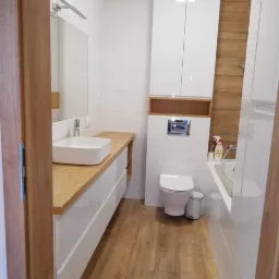 Nowoczesna łazienka z białymi szafkami na wysoki połysk, drewnianym blatem pod umywalką i drewnopodobnymi płytkami na podłodze i ścianie za wanną, widziana z perspektywy wejścia.