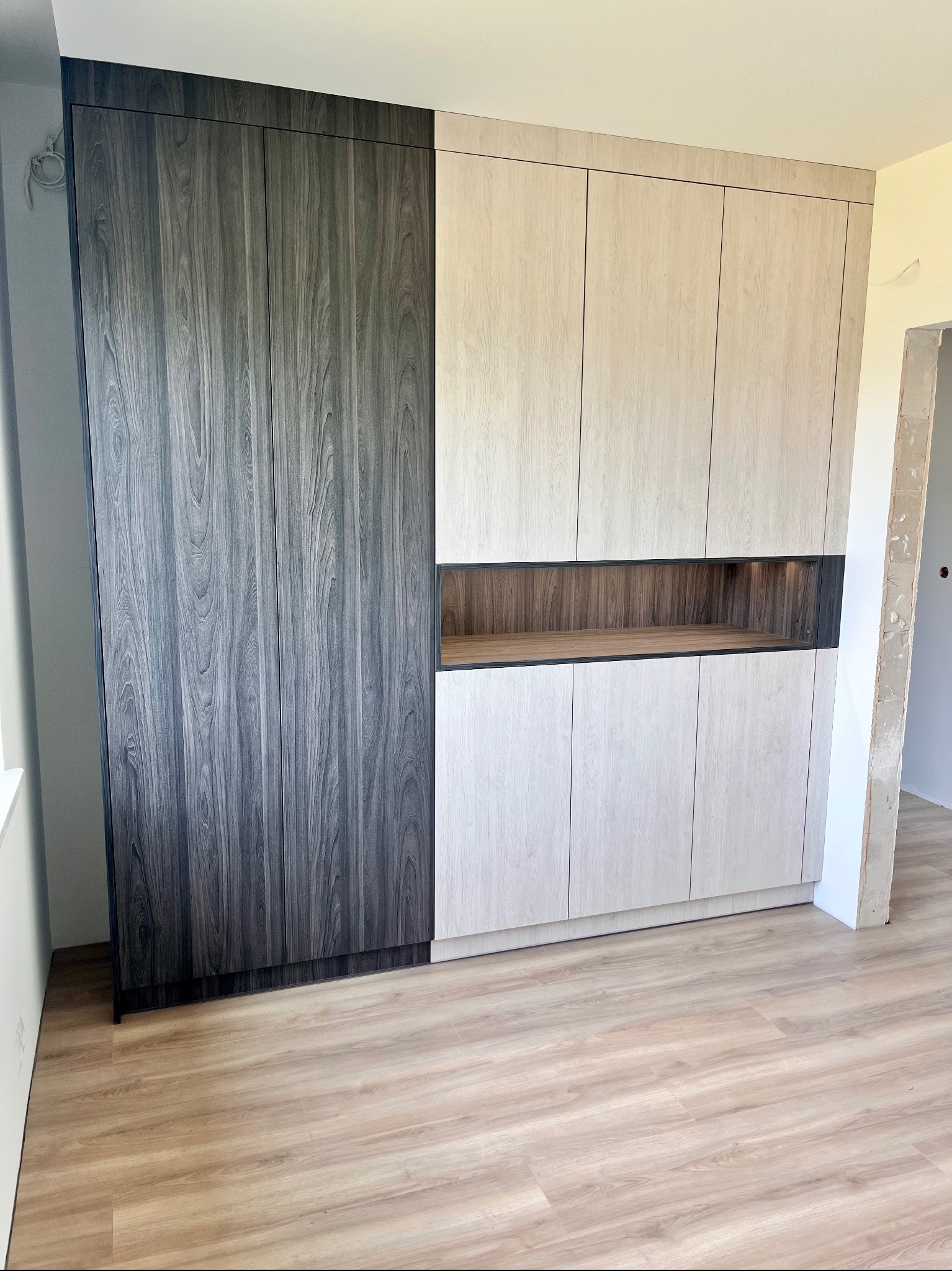 Dwukolorowa szafa na wymiar z drewnianymi frontami, z otwartą półką pośrodku, na tle jasnej podłogi i surowego otworu drzwiowego.