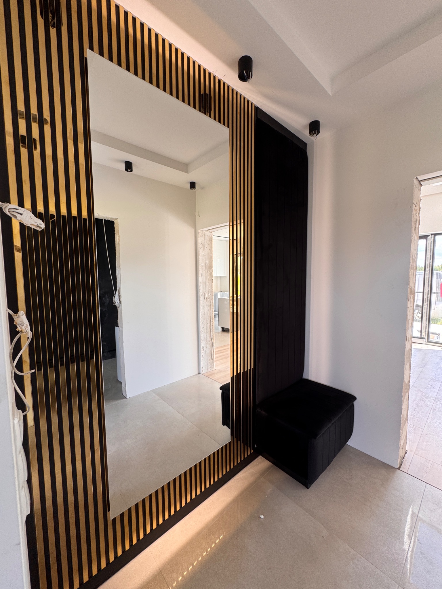 Nowoczesna aranżacja przedpokoju z lustrem w dekoracyjnej ramie z lameli, tapicerowanym siedziskiem i oświetleniem punktowym. Jasna podłoga i białe ściany.