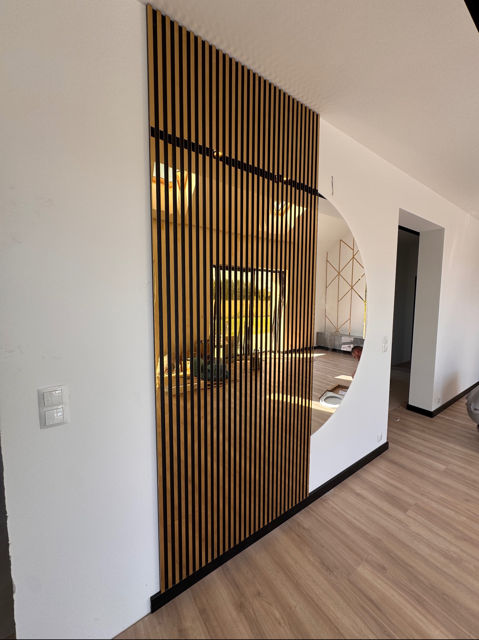 Nowoczesna ściana z drewnianych lameli w kolorze jasnego drewna, częściowo zasłaniająca lustro w kształcie półkola. Widoczna podłoga z paneli i fragment wnętrza.