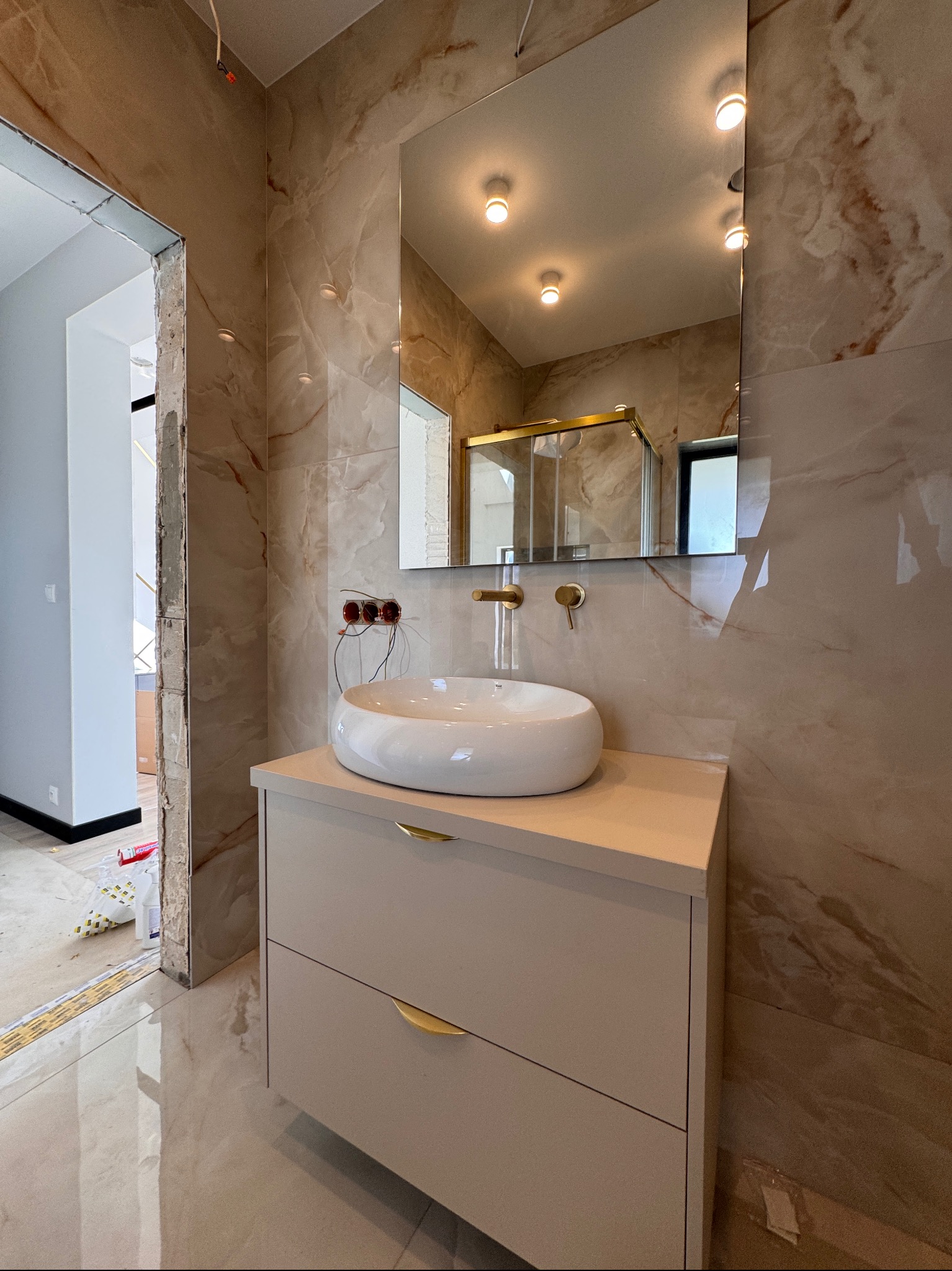 Łazienka w trakcie wykończenia: szafka z umywalką nablatową, lustro, płytki imitujące marmur. Widoczne niedokończone obramowanie drzwi i wystające kable.