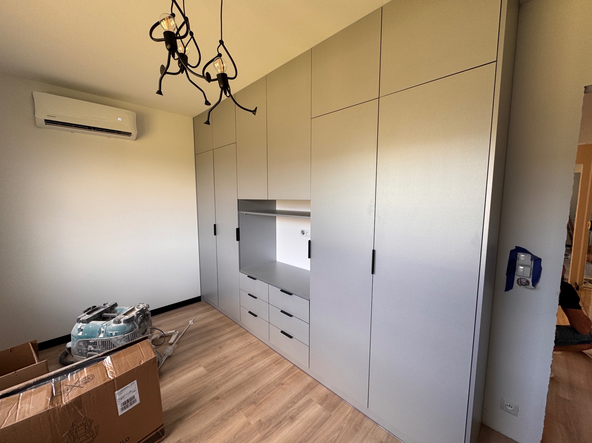 Jasnoszara szafa na wymiar z szufladami i otwartą półką, oświetlona designerską lampą. Podłoga drewniana, widoczne narzędzia i kartony po montażu.