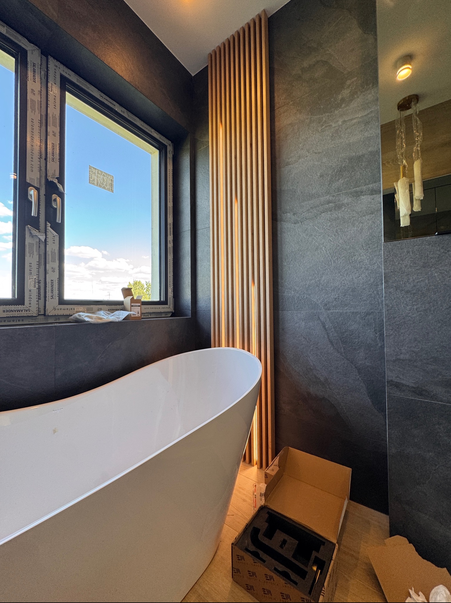Nowoczesna łazienka z wolnostojącą wanną, drewnianą ścianką działową i oknem. Szare płytki na ścianach, pudełka na podłodze. Widok na zewnątrz przez okno.