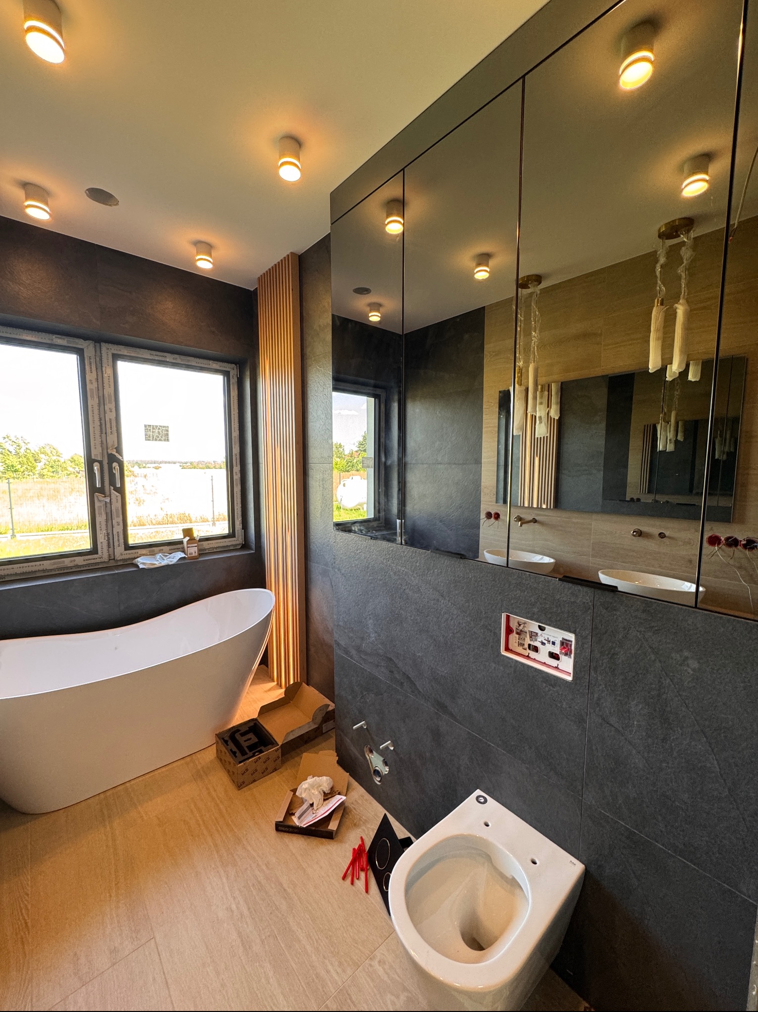 Nowoczesna łazienka w trakcie montażu: wolnostojąca wanna, umywalki w lustrzanej szafce, podwieszana toaleta. Elementy instalacji widoczne na tle szarych płytek.