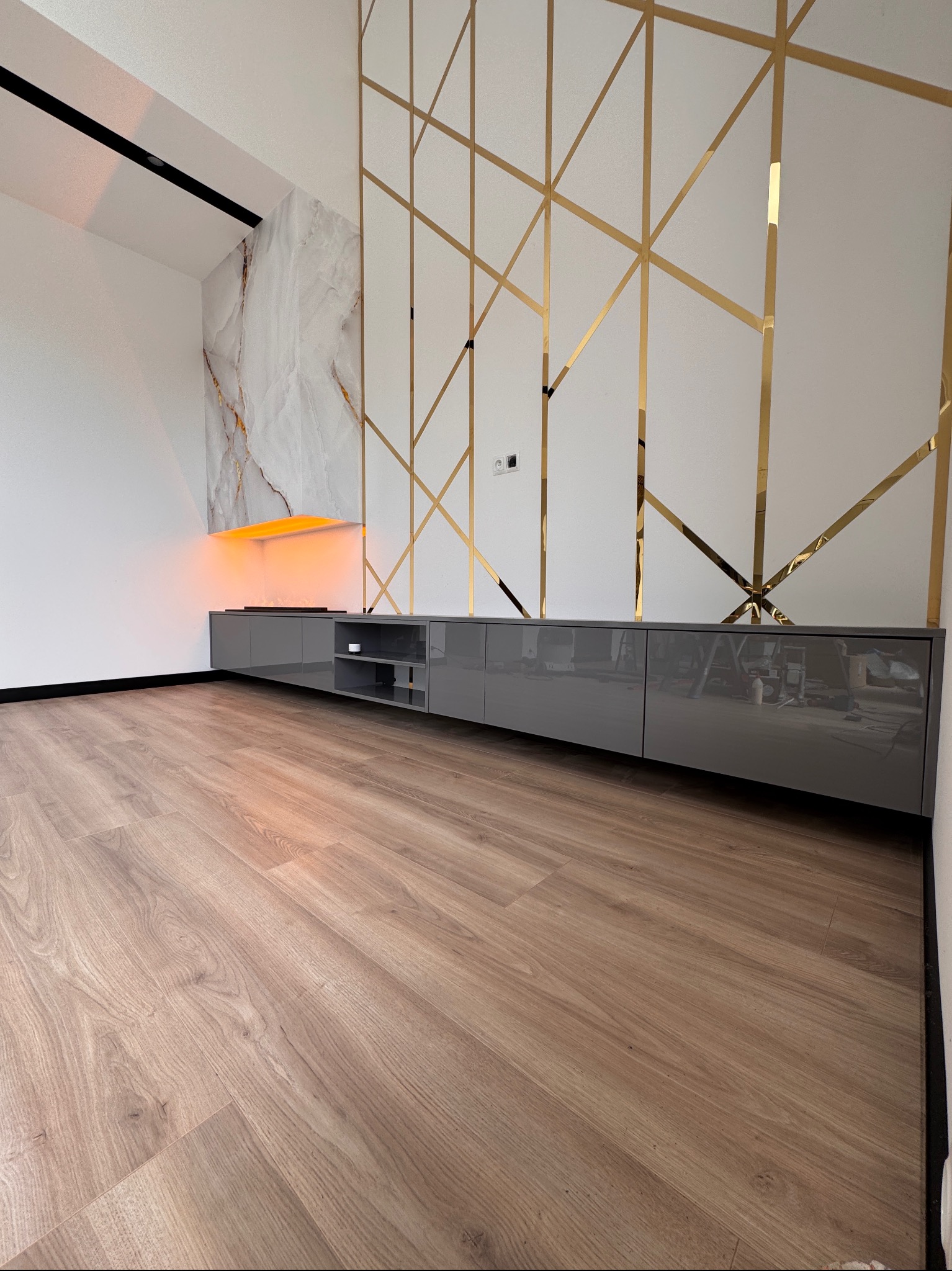 Szara szafka RTV na wymiar z połyskiem, podświetlana od góry pomarańczowym światłem, na tle białej ściany z dekoracyjnymi, złotymi elementami.