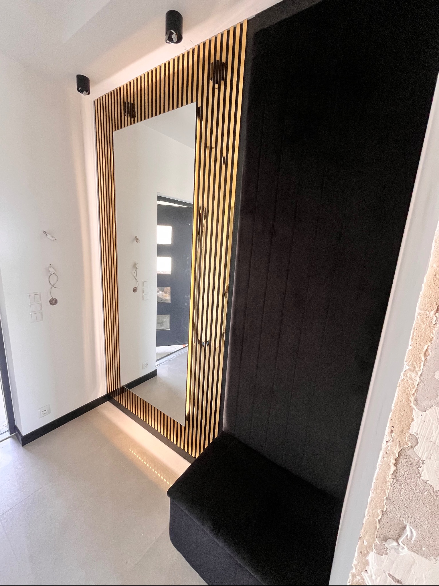 Nowoczesna aranżacja przedpokoju z lustrem w ramie z drewnianych listew i tapicerowanym siedziskiem w kolorze czarnym. Minimalistyczny design, jasne płytki podłogowe.