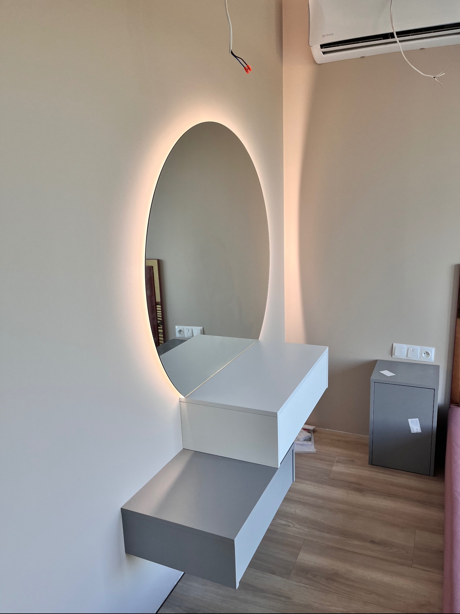 Nowoczesna toaletka z owalnym lustrem podświetlanym LED, zamontowana na ścianie w sypialni. Białe blaty tworzą minimalistyczny design, a drewniana podłoga dodaje ciepła.