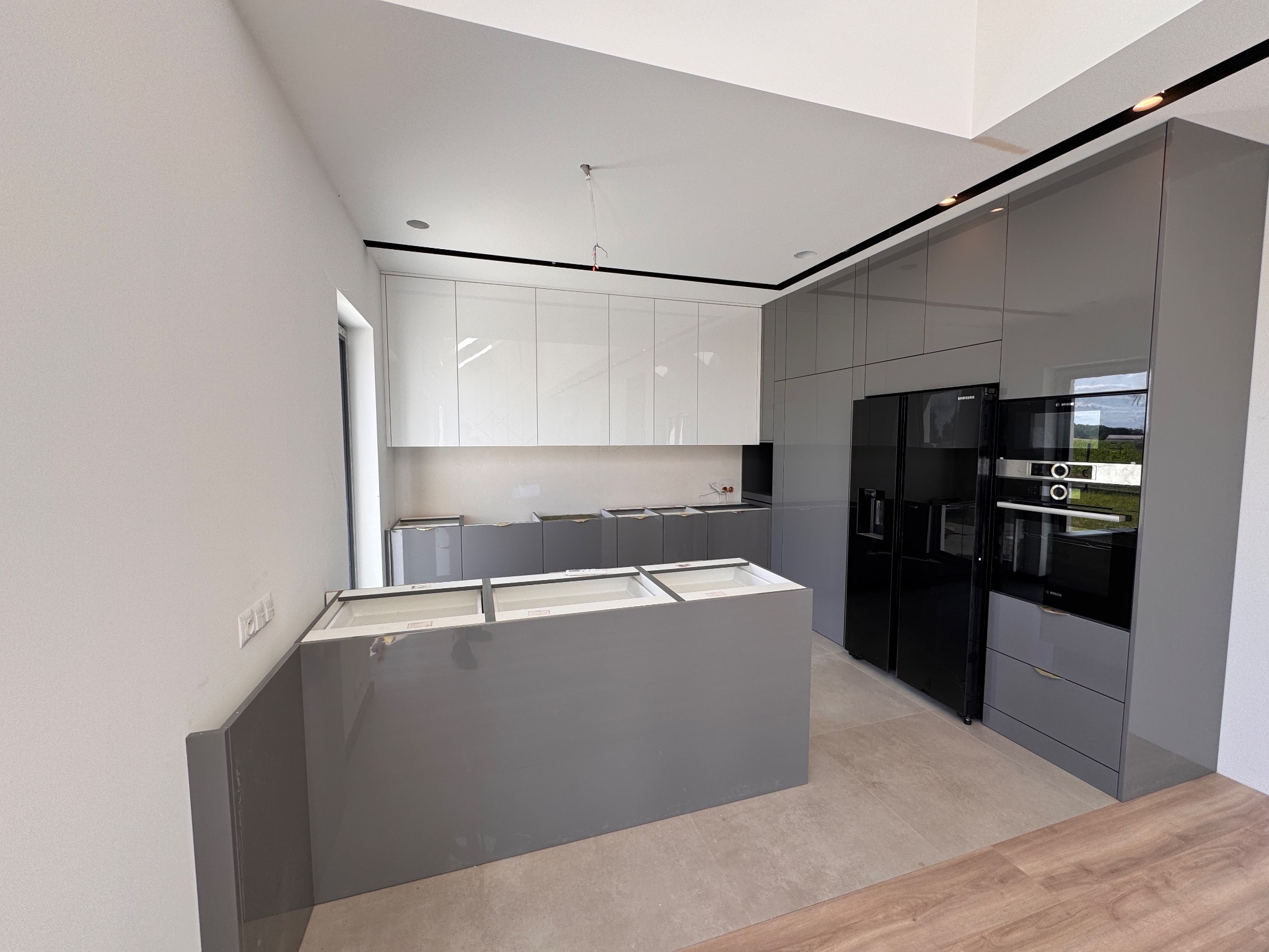 Wnętrze nowoczesnej kuchni w odcieniach szarości i bieli, z wyspą kuchenną w centralnej części i sprzętami AGD marki Samsung i Bosch. Widok z perspektywy szerokokątnej.