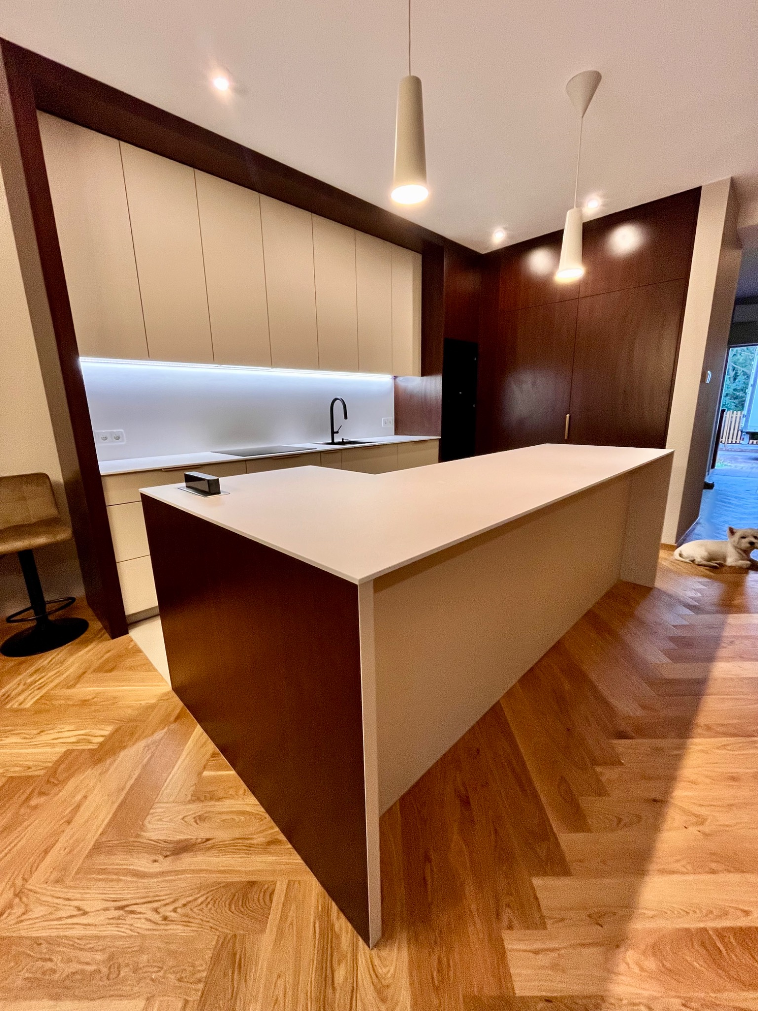 Nowoczesna kuchnia z wyspą, drewniane fronty szafek górnych i dolnych w odcieniach beżu i brązu. Parkiet w jodełkę. Pies leży na podłodze w tle.