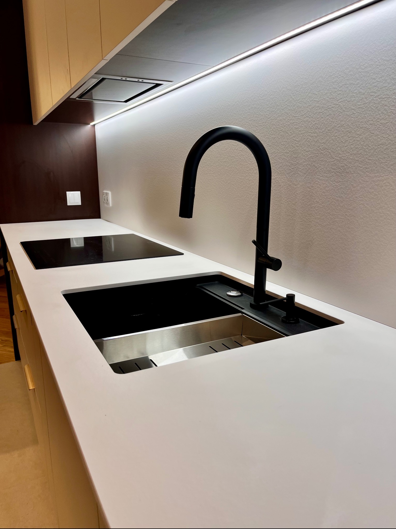 Biały blat kuchenny z czarną armaturą, zlewem i płytą indukcyjną, oświetlony listwą LED. Minimalistyczny design z elementami drewna w tle.