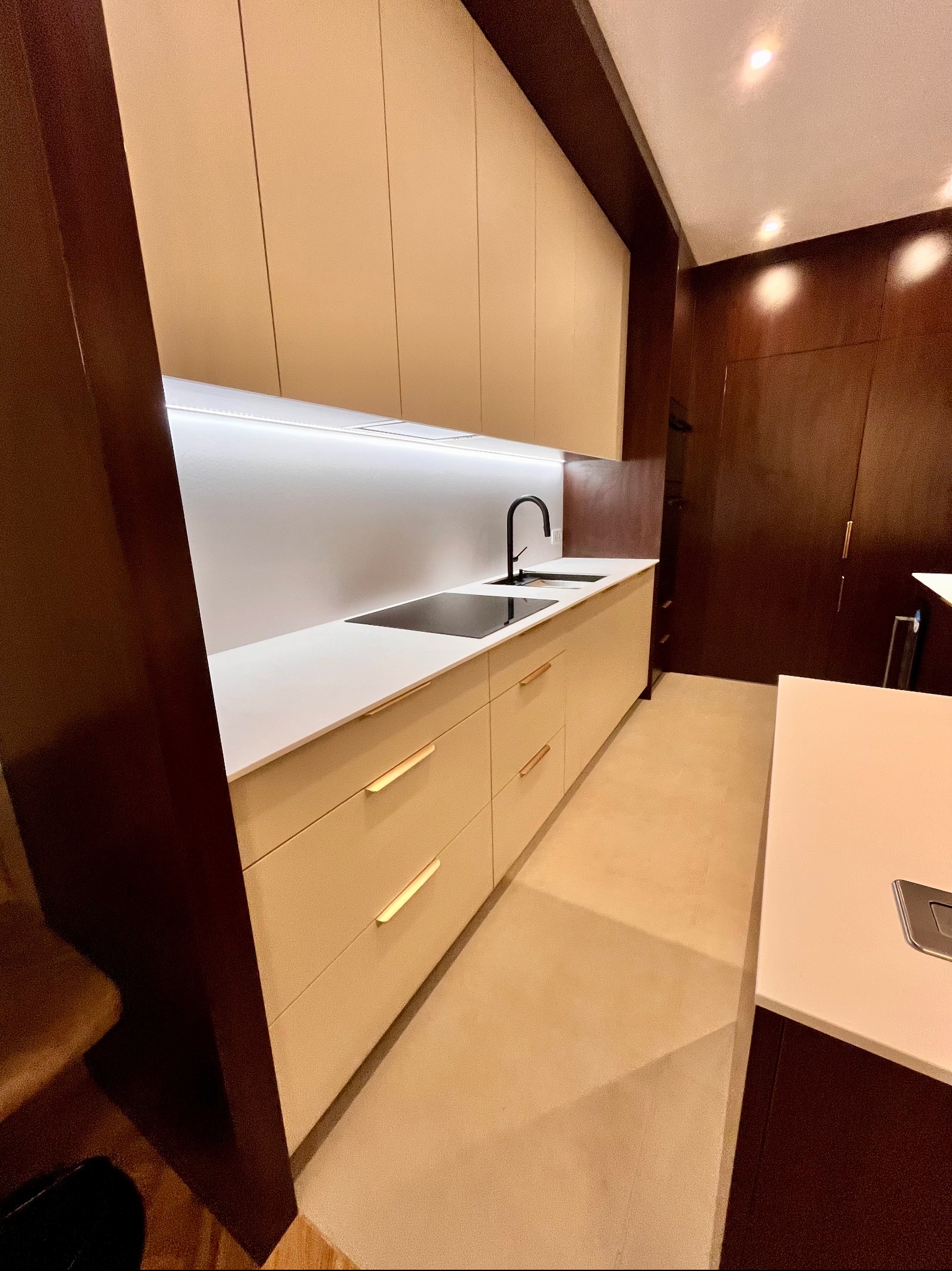 Kremowa kuchnia z matowymi frontami i złotymi uchwytami, oświetleniem LED pod szafkami i czarną armaturą. Ciemne, drewniane wykończenie ścian i wyspa kuchenna.