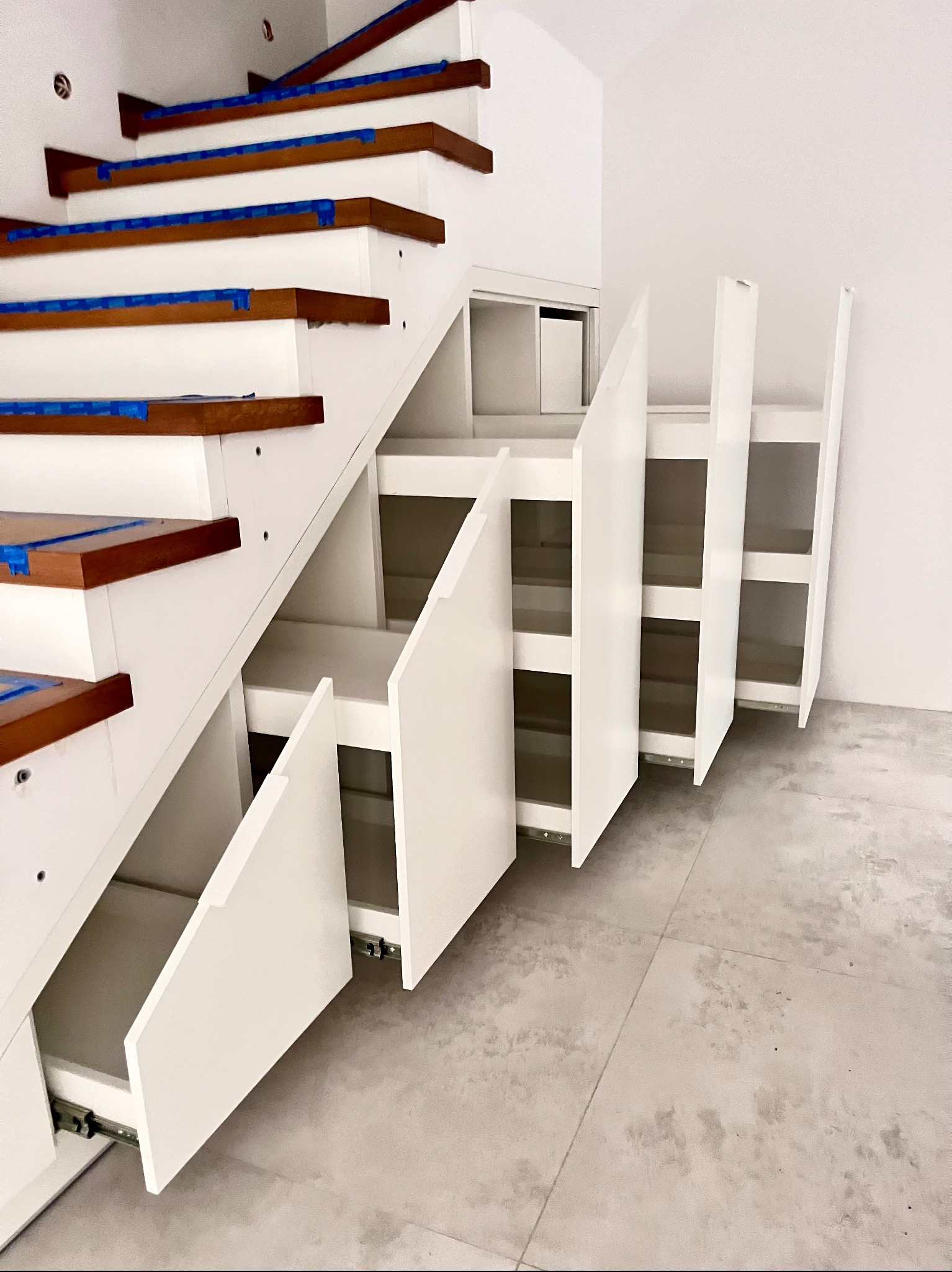 Białe schody z drewnianymi stopniami i wbudowanymi szufladami, częściowo otwarte, ukazując mechanizm wysuwania. Nowoczesny design, minimalistyczne wnętrze.