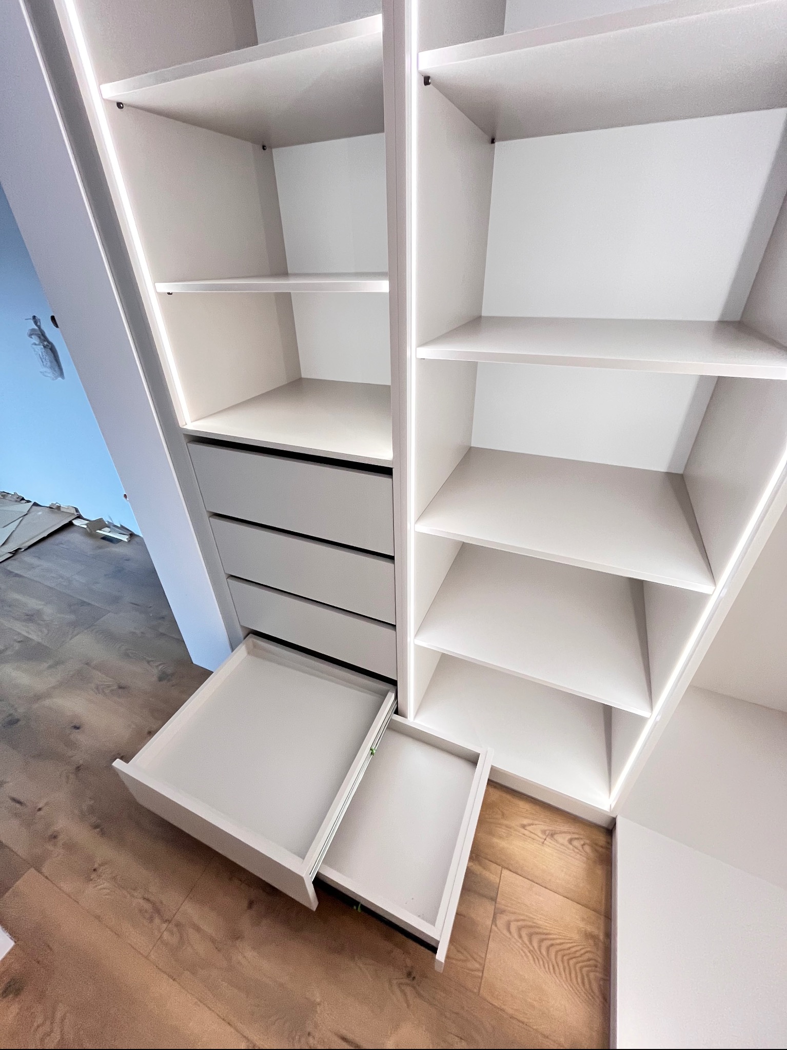 Biała szafa na wymiar ze szufladami i półkami, oświetlenie LED wewnątrz, widok z dołu na drewnianą podłogę. Jedna szuflada wysunięta.