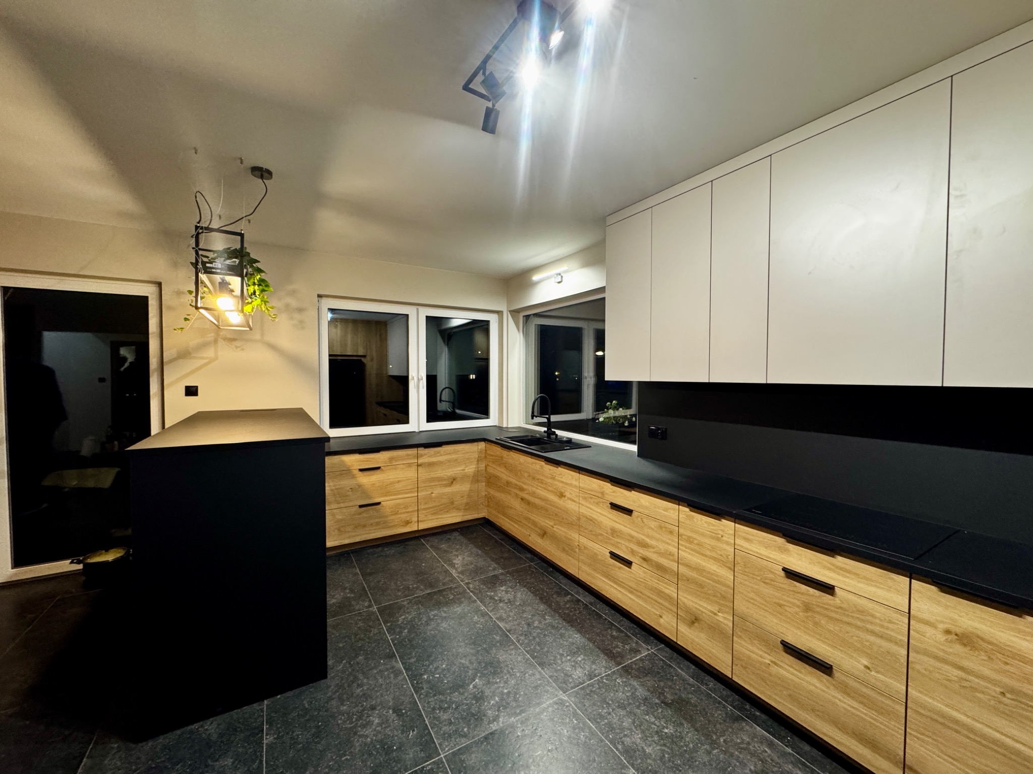Nowoczesna kuchnia z drewnianymi frontami szafek, czarnym blatem i wyspą, oświetlona lampą wiszącą z roślinami. Minimalistyczny design.