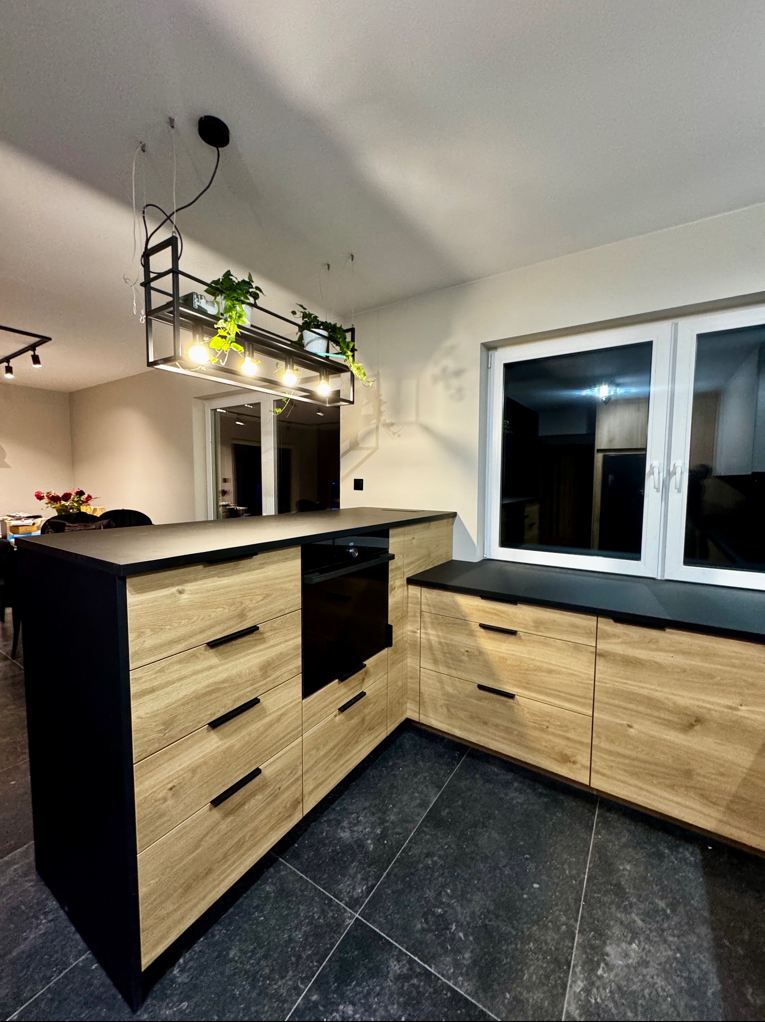 Nowoczesna kuchnia z wyspą: drewniane fronty, czarny blat i detale, oświetlenie z roślinami. Widok na okno i dalszą część domu. Ciemna podłoga z płytek.