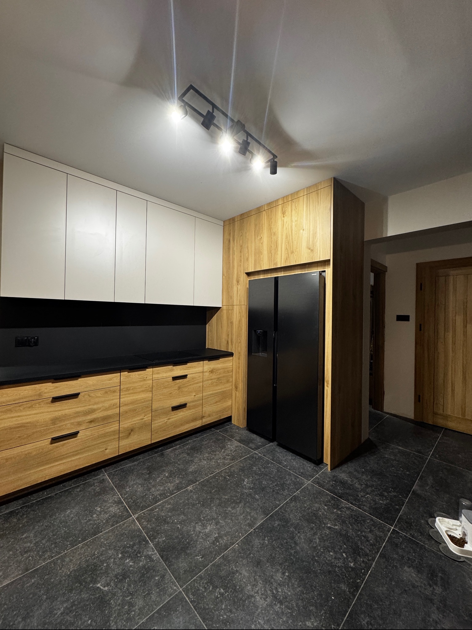 Nowoczesna kuchnia z drewnianymi frontami, białymi szafkami wiszącymi i czarnym blatem. Czarna lodówka side-by-side w zabudowie. Podłoga z ciemnych płyt.