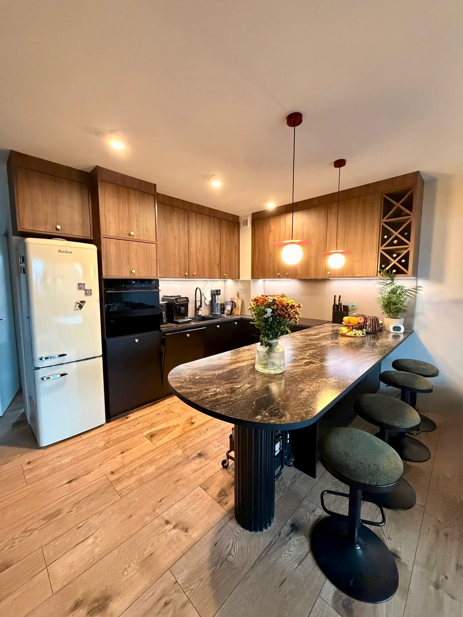 Nowoczesna kuchnia z drewnianymi szafkami, czarnym blatem i barkiem z hokerami. Uwagę przykuwa retro lodówka Amica i designerskie lampy wiszące.