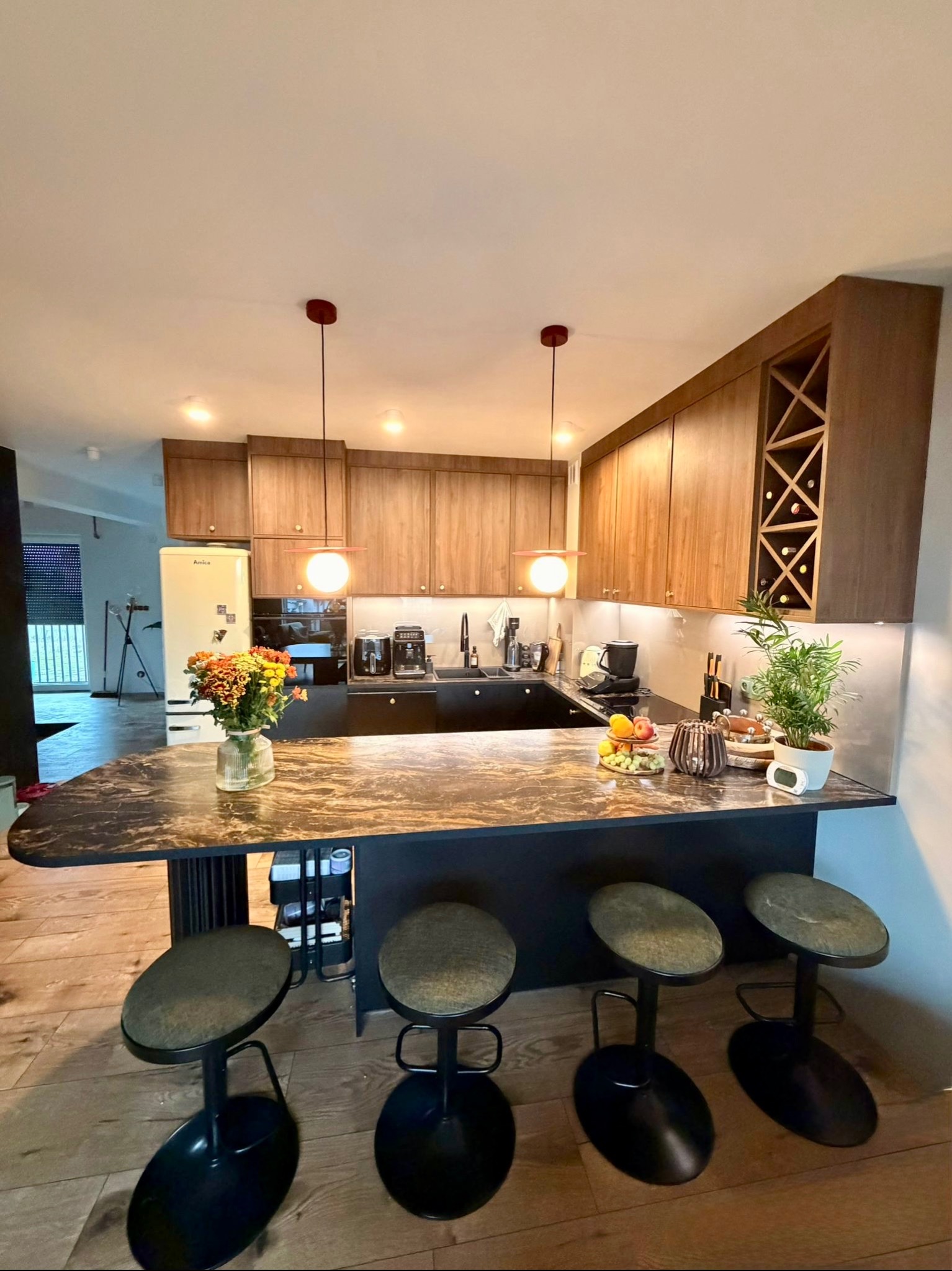 Nowoczesna kuchnia z wyspą i drewnianymi szafkami. Na blacie kwiaty, owoce i dekoracje. Nad wyspą wiszą lampy z czerwonymi akcentami. Widoczne cztery hokery.