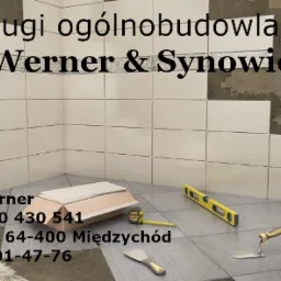 Układanie płytek ceramicznych na ścianie i podłodze w łazience, widoczne narzędzia: poziomica, paca, miarka. Częściowo wykończona ściana, surowy beton w tle.