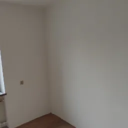 Świeżo pomalowane na biało ściany pokoju z oknem, przez które widać budynek i drzewo, z grzejnikiem pod oknem i butelką wody na parapecie.