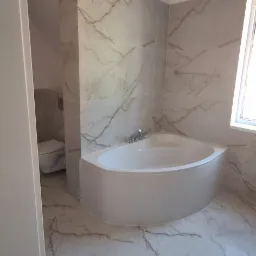 Wykończona łazienka w odcieniach bieli i szarości, z narożną wanną i toaletą w tle, marmurowe płytki na ścianach i podłodze, widok z perspektywy wejścia.