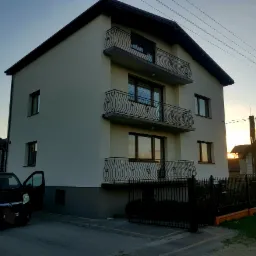 Trzykondygnacyjny dom z ocieploną elewacją w odcieniach beżu, z balkonami o zdobionych, kutych balustradach, widok z boku z otwartymi drzwiami samochodu na podjeździe, zachodzące słońce w tle.