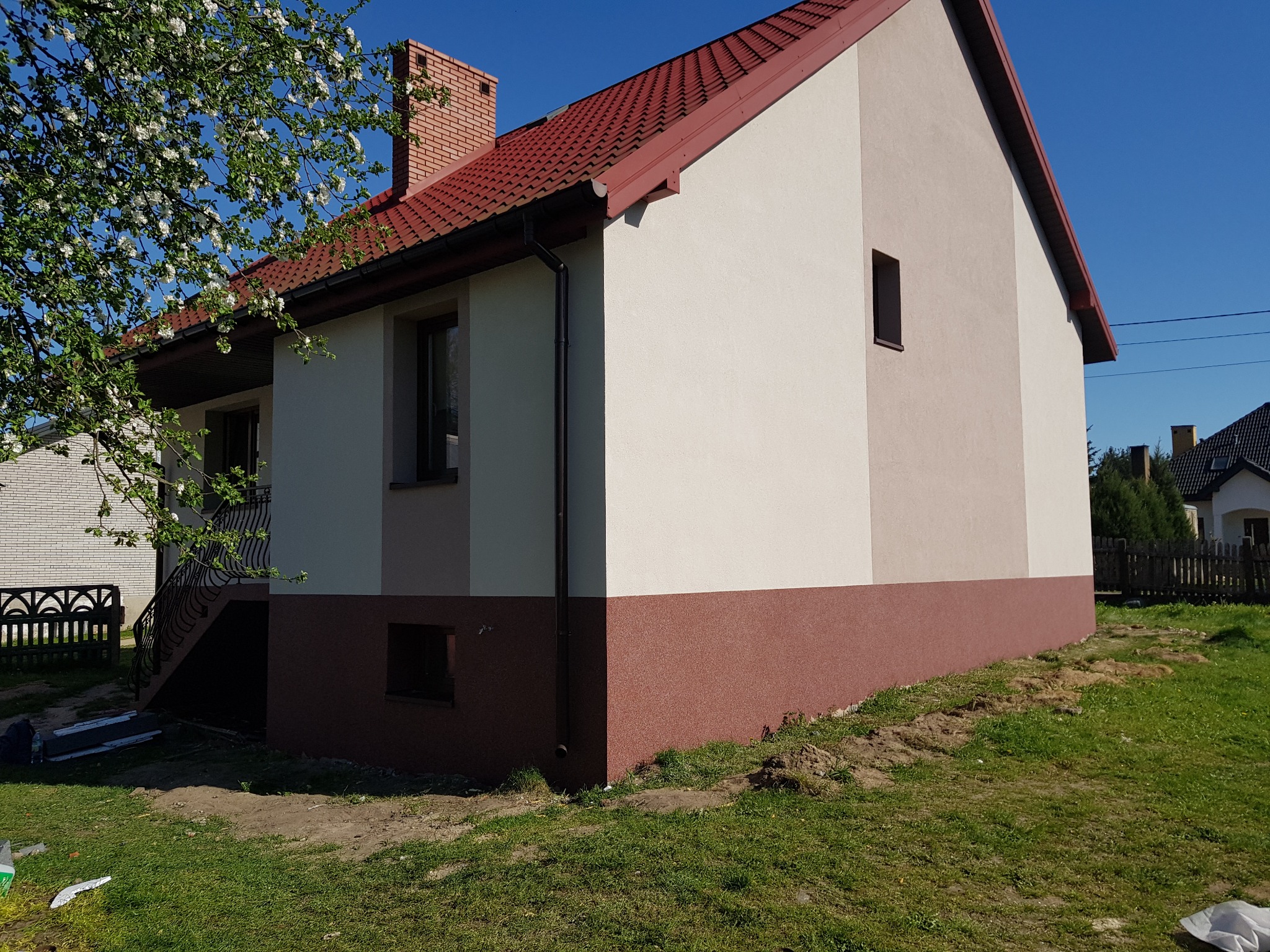 Dwukolorowa elewacja domu jednorodzinnego z czerwoną dachówką, widok z boku na tle kwitnącego drzewa i błękitnego nieba.