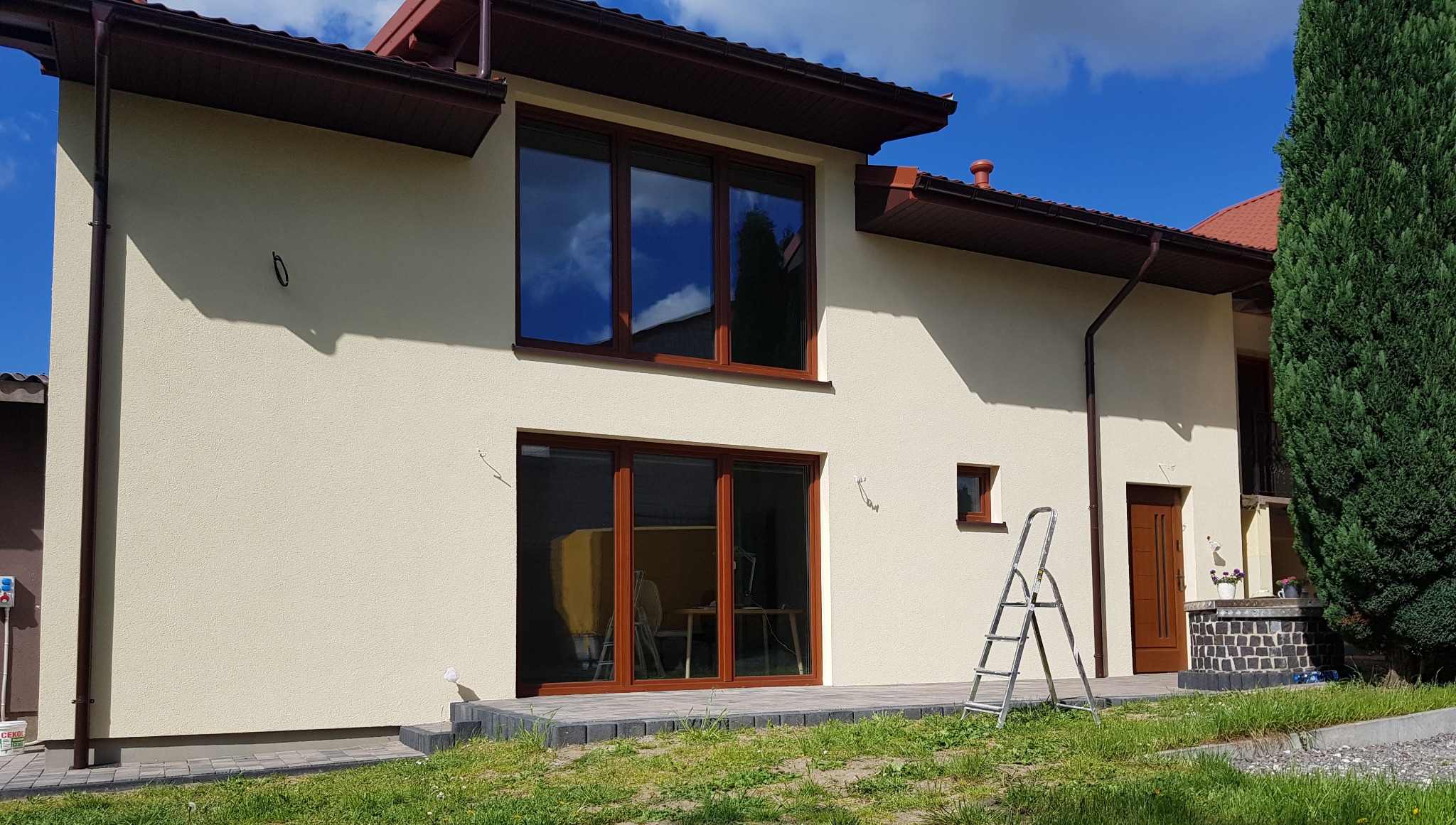 Jasna elewacja domu jednorodzinnego z brązowymi oknami i dachem, widoczna drabina oparta o ścianę, trawnik z przodu.