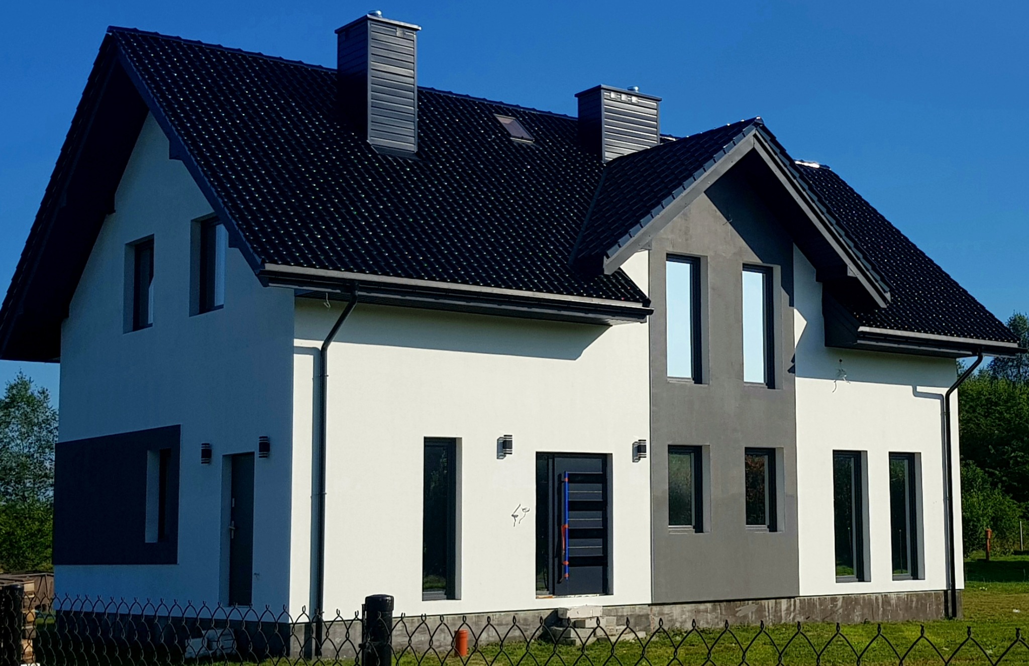 Nowoczesny dom jednorodzinny z biało-szarą elewacją, czarnym dachem i trzema kominami, widoczny zza metalowego ogrodzenia.