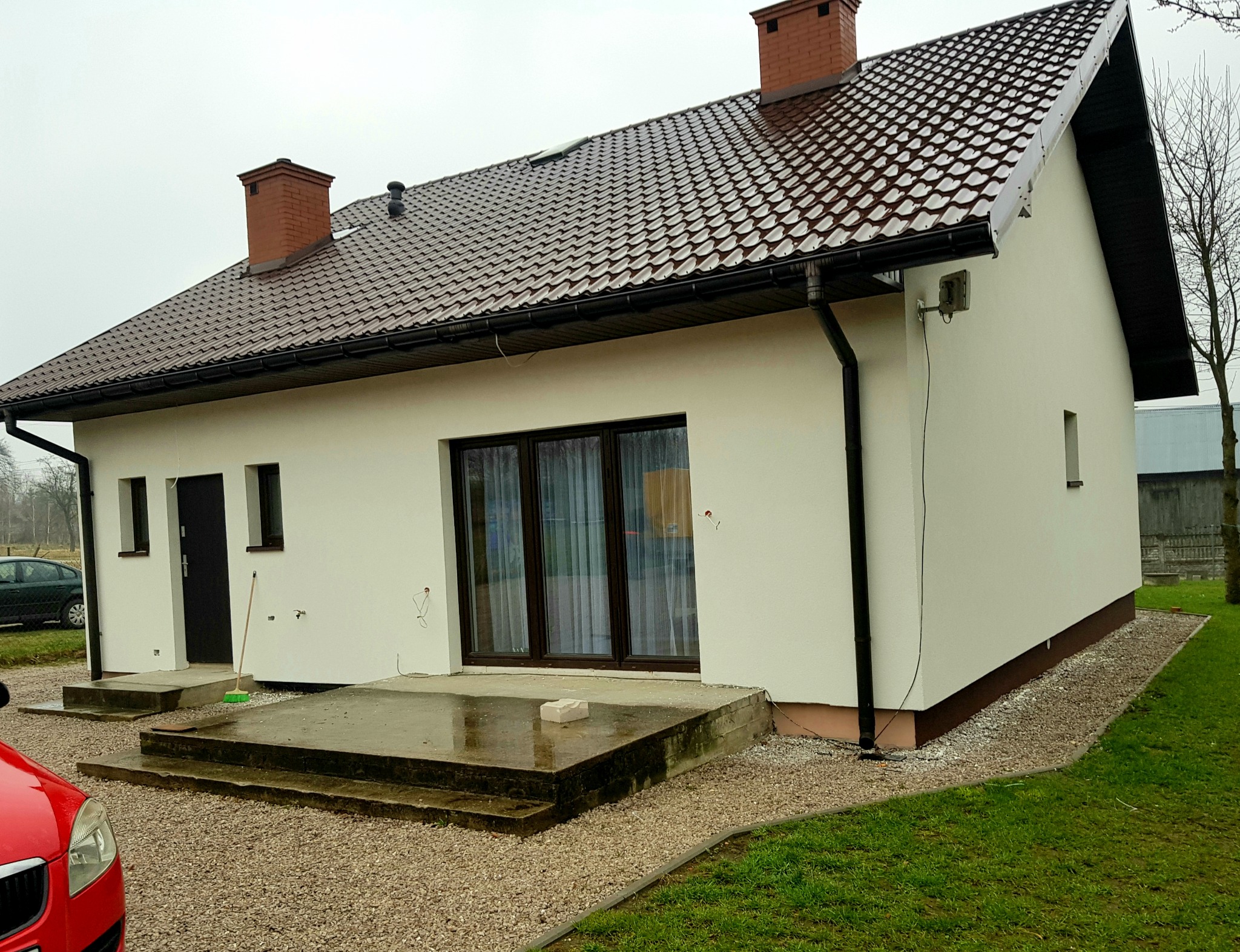 Jednopiętrowy dom z białymi ścianami i brązowym dachem z dachówki, widoczne przygotowania do wykończenia elewacji, niezabezpieczone kable wystające ze ściany, mokry betonowy taras.