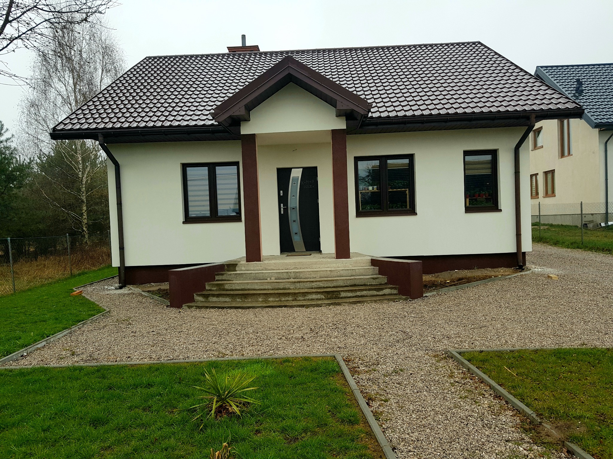 Gotowy, otynkowany dom jednorodzinny z brązowym dachem i stolarką okienną, otoczony żwirowym podjazdem i trawnikiem.