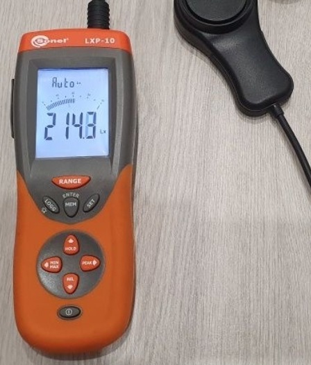 Miernik natężenia oświetlenia Sonel LXP-10 wyświetlający wartość 214.8 lx na podświetlanym ekranie, z czarną sondą światłoczułą obok.