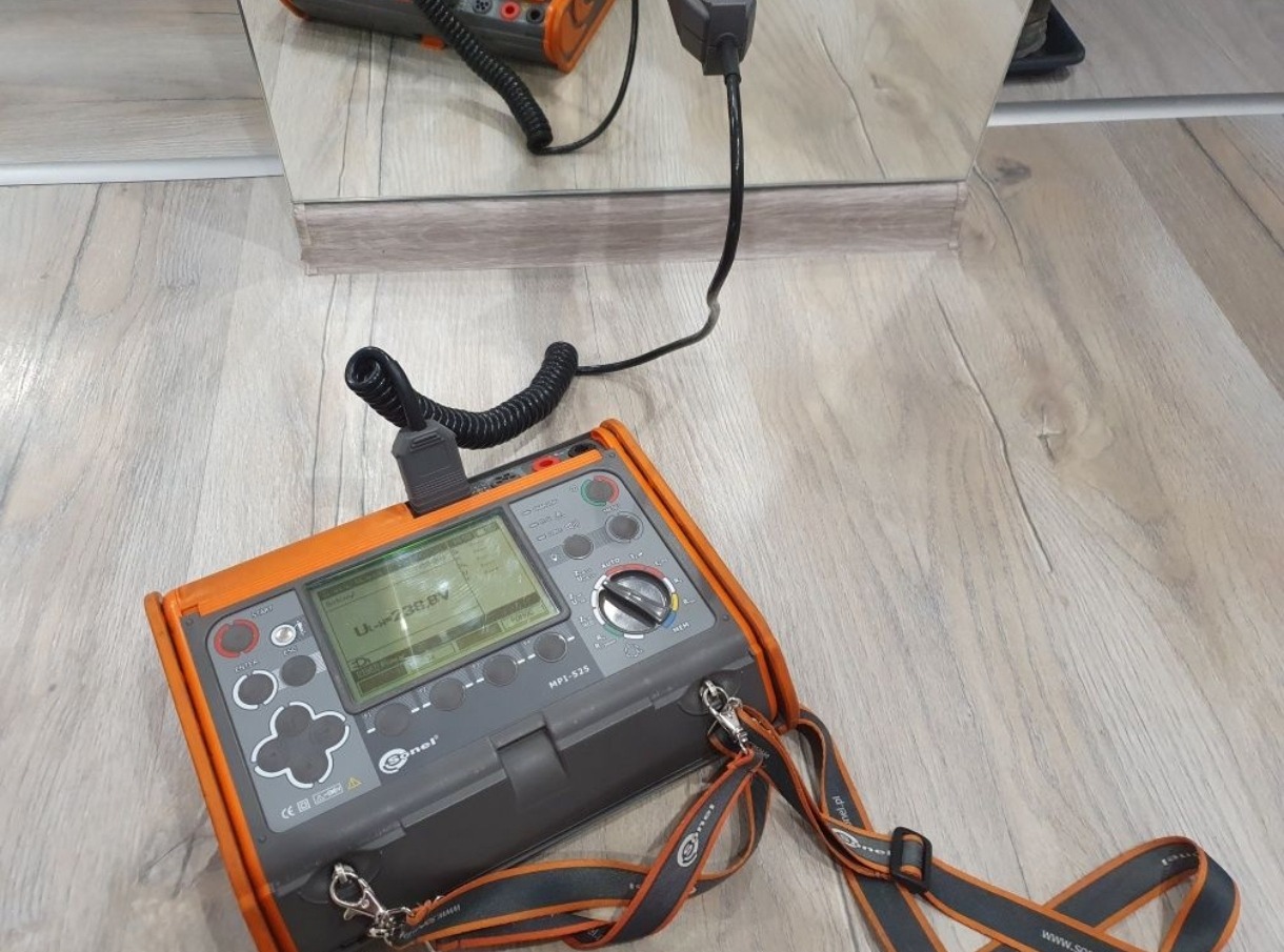 Miernik elektryczny Sonel MPI-525 z podłączonym przewodem, wyświetlający wartość napięcia, leżący na podłodze z paneli, odbicie w lustrze pokazuje gniazdko z wpiętą wtyczką.