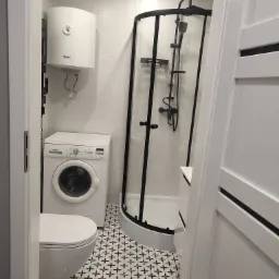 Nowoczesna łazienka z biało-czarną mozaiką na podłodze, kabiną prysznicową z czarnymi ramami, białą pralką Siemens i podgrzewaczem wody, widok zza otwartych białych drzwi.