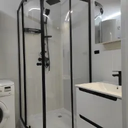 Nowoczesna łazienka z kabiną prysznicową w czarnej ramie, białą umywalką z szafką i pralką, wykończona szarymi płytkami i wzorzystą podłogą.