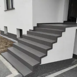 Zewnętrzne schody wejściowe do domu wyłożone szarymi płytkami ceramicznymi z subtelnym wzorem, z wykończonym cokołem budynku w ciemniejszym odcieniu szarości i widocznymi workami z zaprawą w tle.