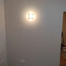 Ściana pomalowana na szaro z kwadratową lampą ścienną i gniazdkiem elektrycznym; szara listwa przypodłogowa i podłoga z płytek w tle widoczny fragment innego pomieszczenia.
