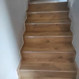 Schody wykończone panelami drewnopodobnymi z metalowymi listwami, widoczne przy szarej ścianie w Malni.