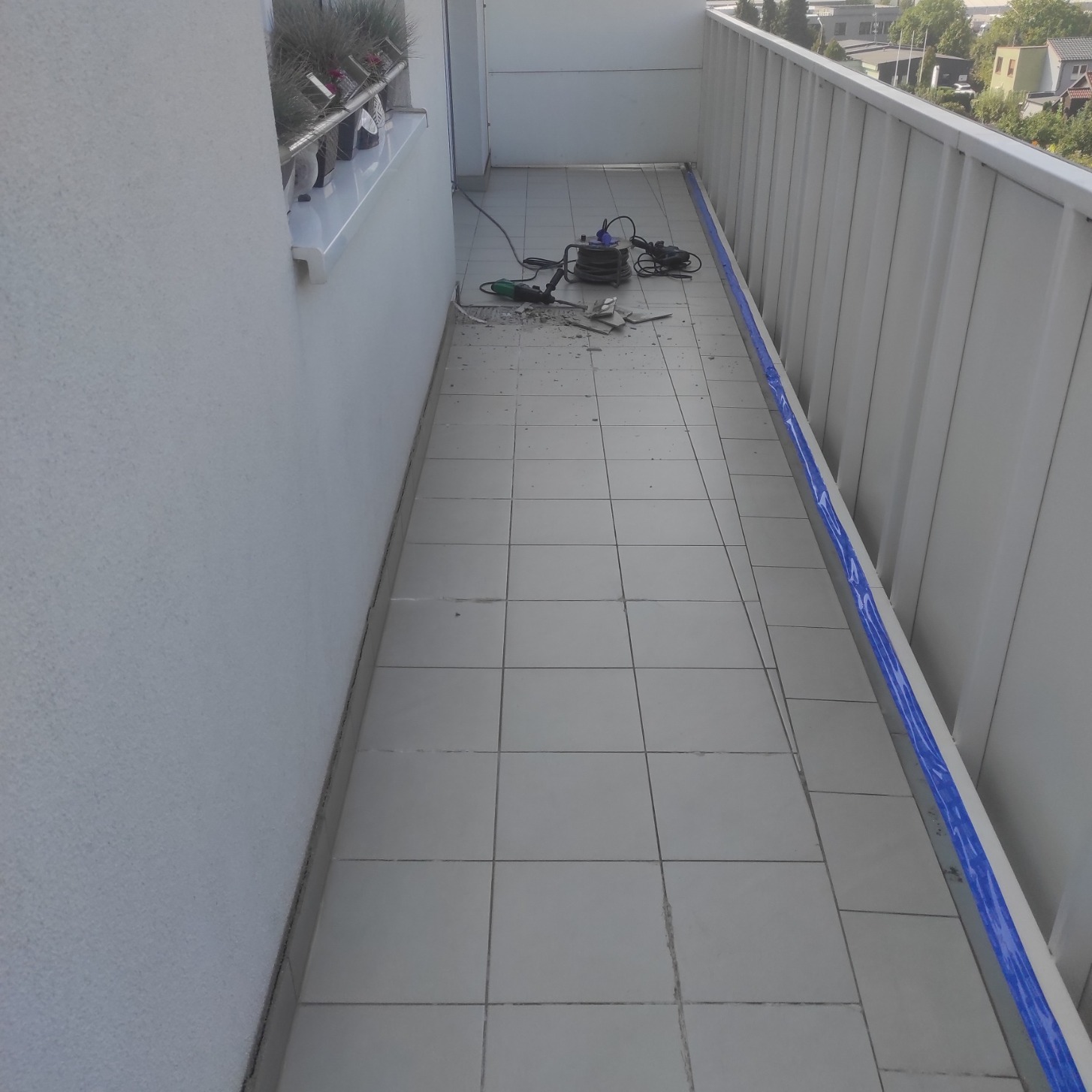Balkon w trakcie prac: zerwane płytki podłogowe, widoczna szlifierka kątowa i odkurzacz przemysłowy, zabezpieczona barierka folią malarską.