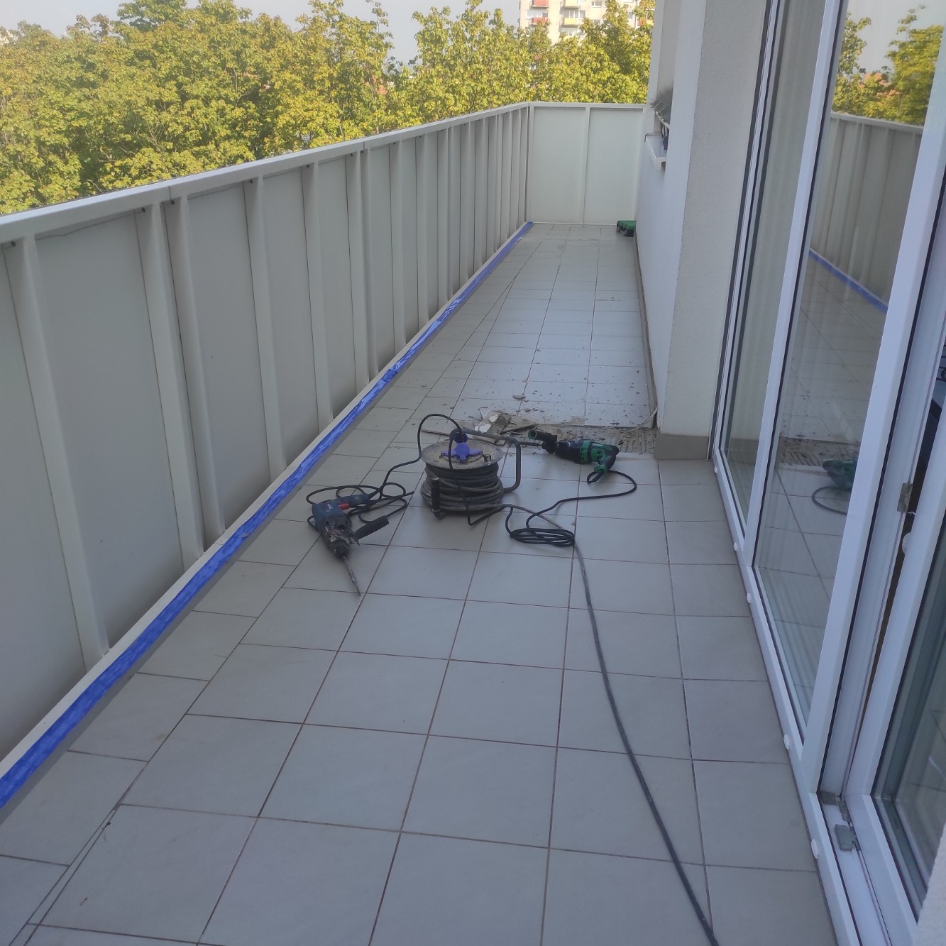Balkon w trakcie remontu: zabezpieczona taśmą balustrada, rozłożone narzędzia (wiertarka, młot udarowy) i przedłużacz na kafelkach.